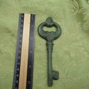 May include: An antique-style, green-colored decorative key with ornate detailing. The key is approximately 8 inches long, with a decorative head and a detailed shaft. A ruler is placed next to the key for scale.