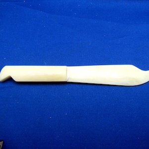 May include: A cream-colored, hand-carved knife with a curved blade and a handle with a notch. The knife is made of a light-colored material and is set against a blue background.