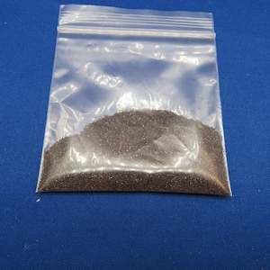May include: A clear plastic bag filled with dark brown granules, possibly sand or a similar substance. The bag is sealed at the top and sits against a solid blue background.
