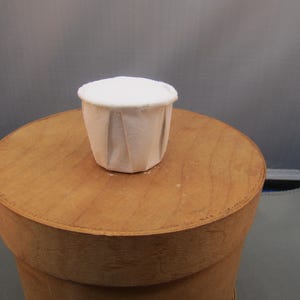 May include: A small, white, paper cup with a pleated design sits atop a round, wooden table. The cup has a slightly textured appearance and a flat, white top. The table is light brown and has a smooth surface.