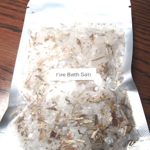 May include: A clear, sealed bag filled with white bath salts and dried herbs. The label reads "Fire Bath Salt". The salts appear coarse and are mixed with brown and green plant matter. The bag is on a wooden surface.