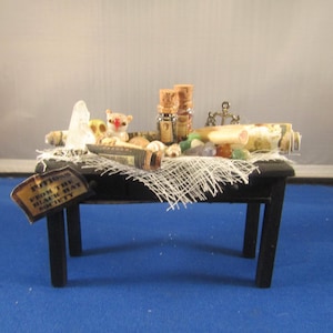 May include: A black wooden table with a white lace tablecloth. The table is decorated with various items, including a small white cat figurine, a crystal, a rolled up scroll, and several small bottles with cork tops. A sign on the table reads "Potions from the Black Hat Society."