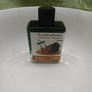 May include: A small, square, dark green glass bottle with a black cap. The label reads "Lodestone Piedra Iman Oil Aceite" with an illustration of a magnet and a rock. The bottle is 0.5 fl oz.