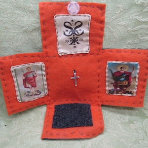May include: A cross-shaped orange felt fabric piece featuring a small silver cross at the center. It includes three rectangular fabric patches: one with a black symbol at the top, and two depicting figures in red and blue robes on the sides.