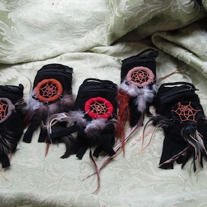 May include: Five black pouches with zipper closures, each adorned with a dreamcatcher design. The dreamcatchers feature intricate web patterns in orange, red, and pink, accented with feathers and beads. The pouches are displayed on a textured, light green surface.