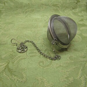 May include: A stainless steel tea infuser with a mesh basket and a chain attached to a pentagram charm. The infuser is resting on a green patterned fabric.