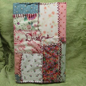 May include: A patchwork journal with a variety of floral and dotted fabric squares in shades of pink, blue, and cream. The word "Dreams" is embroidered in cursive on the front. The edges are stitched with a contrasting dark red thread.