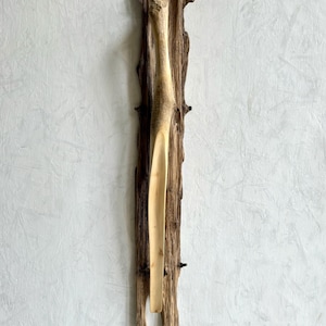 Könnte beinhalten: Eine Holzskulptur einer menschlichen Figur mit einem langen, dünnen Körper und einem runden Kopf. Die Skulptur ist auf einem Stück Treibholz mit einer rauen, natürlichen Textur montiert.