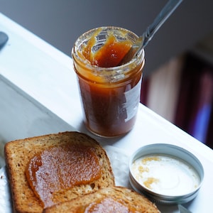 May include: Two slices of toasted bread topped with a thick layer of orange-colored fruit spread. A glass jar of the spread is in the background, with a spoon resting on the rim.