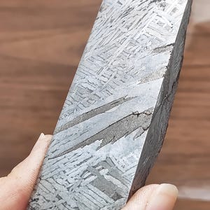 Op de afbeelding: Een gepolijst, rechthoekig meteorietfragment met een Widmanstätten-patroon. Het metallic grijze oppervlak toont een kruisend, geometrisch ontwerp. De randen zijn ruw en donker en onthullen de natuurlijke, ongepolijste textuur van de ruimterots.
