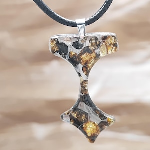 May include: A pendant necklace with a distinctive hammer-shaped design. The pendant is crafted from clear resin, incorporating silver and golden-brown stones. It is suspended from a black cord with a silver clasp, offering a striking visual contrast.