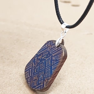 May include: A rectangular pendant with a distinctive patterned design in shades of blue and brown, suspended from a black cord necklace. The pendant is attached to a silver-coloured bail.