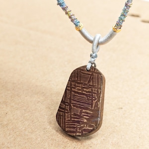 May include: A brown pendant necklace with a geometric pattern. The pendant is rectangular with rounded edges and hangs from a grey cord with colourful accents. The necklace has gold-coloured beads.