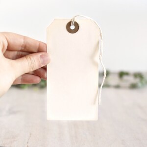 Puede incluir: Una etiqueta blanca en blanco con un ojal de metal y una cuerda atada. La etiqueta est&aacute; sostenida en una mano.