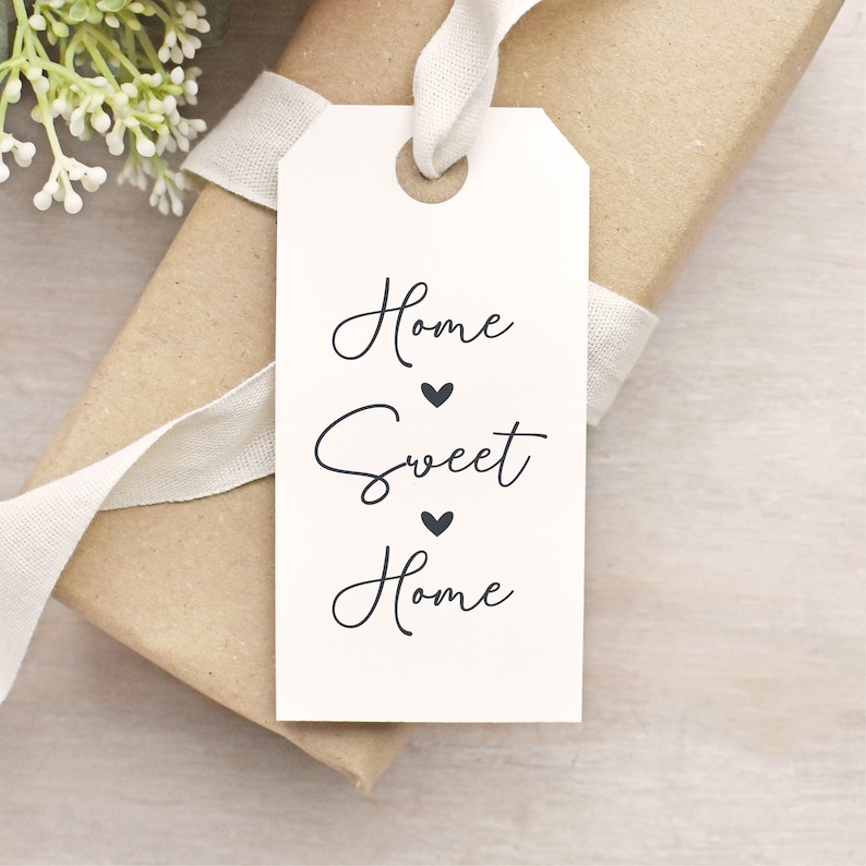 May include: A gift tag with the words "Home Sweet Home" in elegant script, with heart symbols. The tag is white and attached to a wrapped package with a cream ribbon. A sprig of white flowers is in the background.