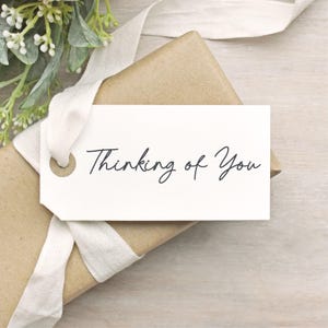 Könnte beinhalten: Ein rechteckiges weißes Geschenkanhänger-Schild mit dem schwarzen Aufdruck Thinking of You, befestigt mit einem weißen Stoffband an einem braunen, in Papier eingewickelten Paket, begleitet von einem kleinen Büschel grüner Blätter mit weißen Blütenknospen.