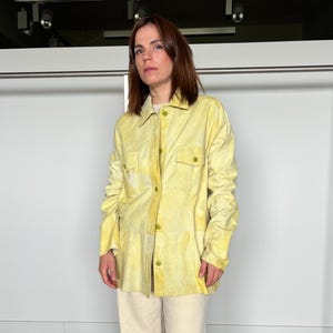 Peut inclure: Une personne porte une chemise jaune boutonnée avec deux poches de poitrine et un pantalon clair. La chemise a un aspect délavé. Le pantalon est de couleur beige clair. La personne se tient devant un mur blanc.