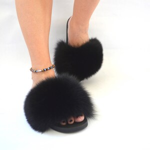 K&ouml;nnte beinhalten: Schwarze Pelz-Sandalen mit flauschigem, dickem Pelz-Futter. Die Sandalen haben einen einfachen schwarzen Riemen und eine schwarze Sohle.
