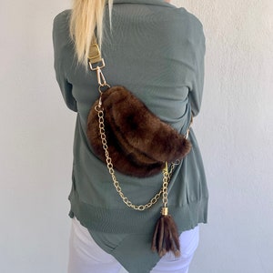 May include: A brown faux fur fanny pack with a gold chain strap and a brown fur tassel. The bag is worn by a person wearing a green sweater and white pants.