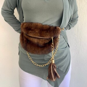 May include: A brown fur waist bag with a gold chain and tassel. The bag is worn over a sage green v-neck top and a matching cardigan.