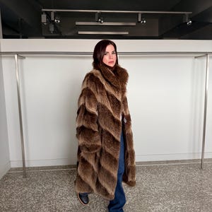 Puede incluir: Un abrigo largo de piel marrón con un diseño en forma de cheurón. El abrigo se lleva con vaqueros azules y zapatos negros. El abrigo tiene un cuello ancho y un cierre de botón. El fondo es una pared blanca y una percha de metal.