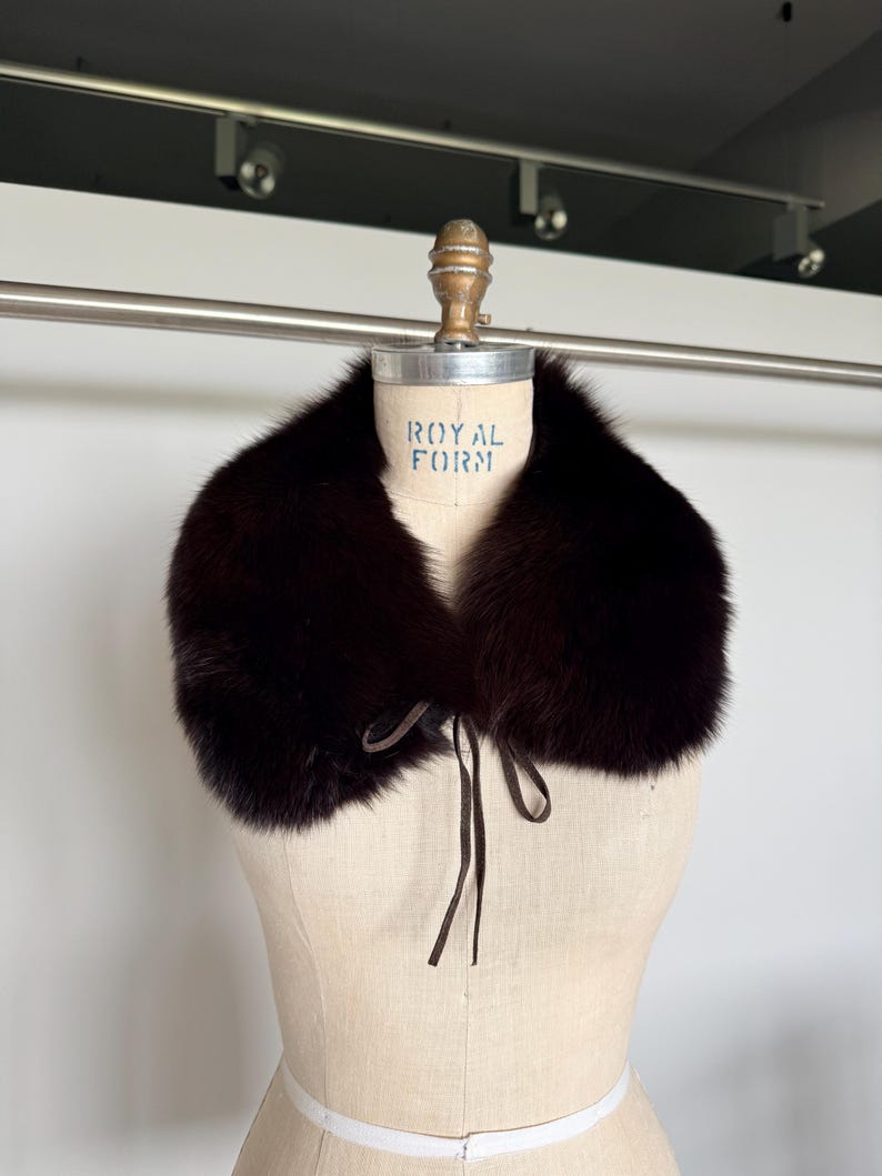 May include: A dark brown fur collar with a tie closure, displayed on a vintage dress form. The collar is plush and appears to be made of fox fur. The dress form has the text "ROYAL FORM" printed on it.