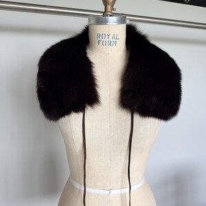 May include: A dark brown fur collar with long, thin ties. The collar is displayed on a mannequin with the text "ROYAL FORM" visible. The fur appears soft and luxurious, perfect for adding warmth and style.