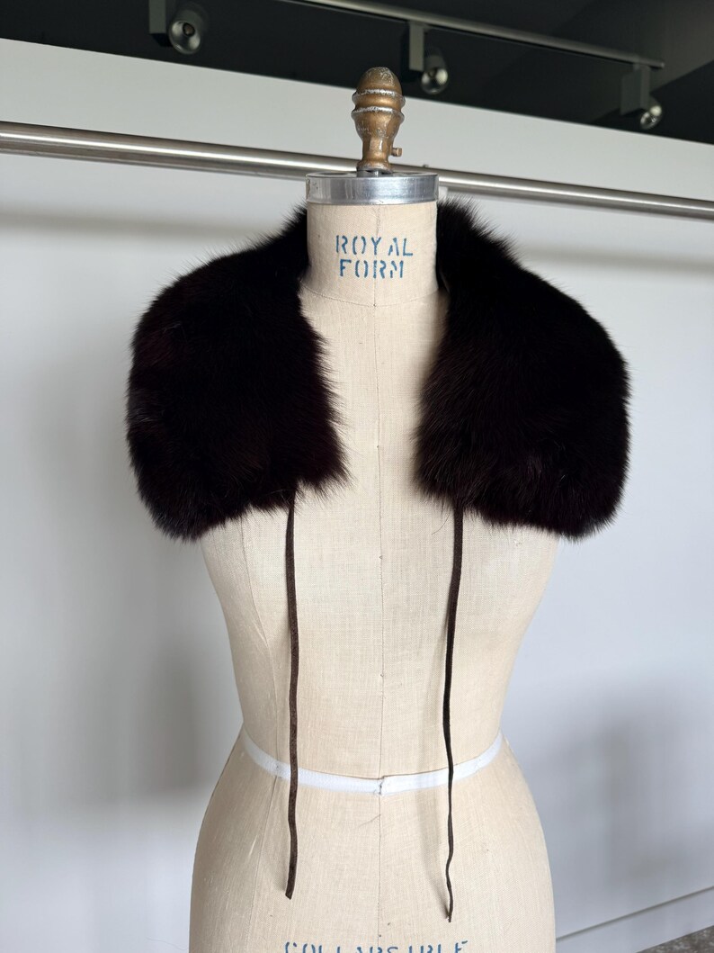 May include: A dark brown fur collar with long, thin ties. The collar is displayed on a mannequin with the text "ROYAL FORM" visible. The fur appears soft and luxurious, perfect for adding warmth and style.