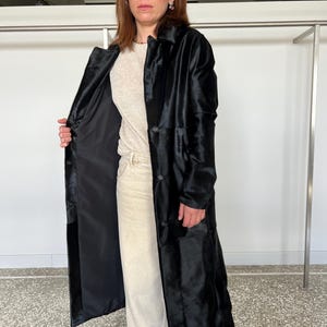 May include: A long, black coat with a textured, fur-like appearance. The coat has a button-down front and a classic collar. The interior lining is black. The person is wearing a light-colored top and pants. The coat appears to be knee-length.