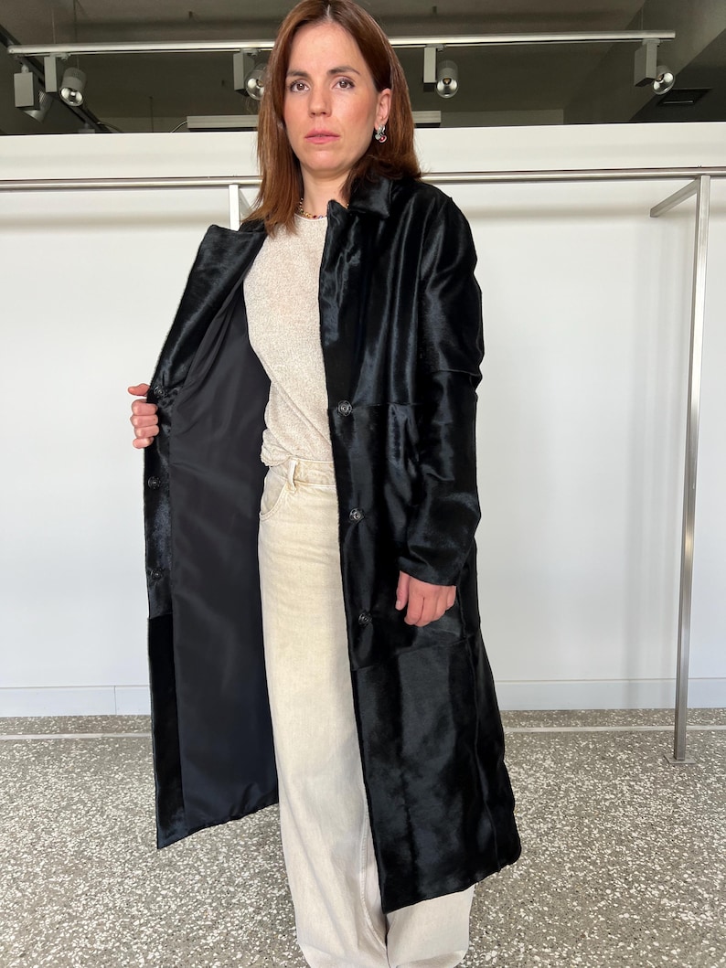 May include: A long, black coat with a textured, fur-like appearance. The coat has a button-down front and a classic collar. The interior lining is black. The person is wearing a light-colored top and pants. The coat appears to be knee-length.