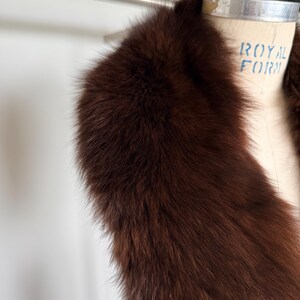 May include: A luxurious, dark brown fur stole. The fur is thick and plush, with a rich, warm tone. The stole is draped over a vintage dress form, showcasing its shape and texture. The background is a soft, neutral color.