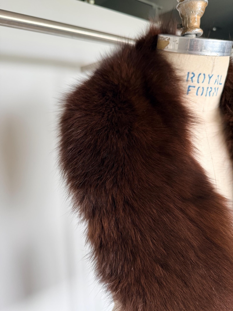 May include: A luxurious, dark brown fur stole. The fur is thick and plush, with a rich, warm tone. The stole is draped over a vintage dress form, showcasing its shape and texture. The background is a soft, neutral color.