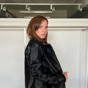 May include: A black coat with a textured surface. The coat has long sleeves and a classic collar. The person in the image is wearing the coat. The background is a white wall with a metal bar.