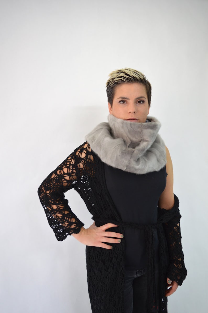 May include: A woman wearing a black crochet cardigan over a black top and jeans. She is wearing a grey fur scarf around her neck.