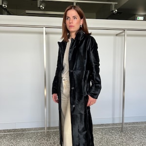 May include: A long, black coat with a button-up front. The coat is knee-length and has a textured appearance. The person is wearing a light-colored sweater and pants. The background is a neutral white wall and a metal clothing rack.