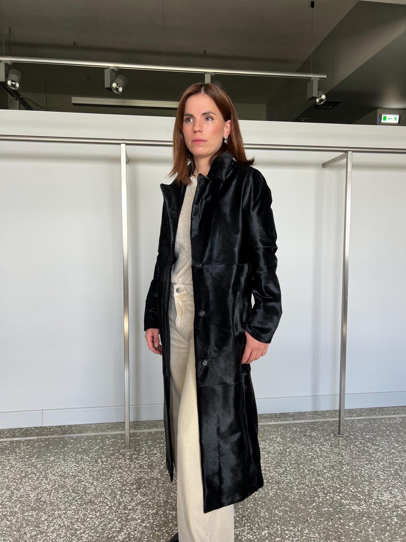 May include: A long, black coat with a button-up front. The coat is knee-length and has a textured appearance. The person is wearing a light-colored sweater and pants. The background is a neutral white wall and a metal clothing rack.