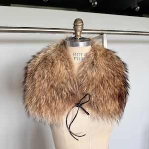 Op de afbeelding: Een luxe bontkraag met bruine en zwarte tinten. De kraag wordt getoond op een mannequin en heeft een donkerbruine striksluiting. De tekst "ROYAL FOR" is zichtbaar op de mannequin.