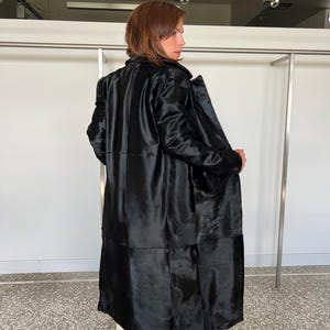 May include: A long, black coat with a shiny, textured appearance. The coat has a tailored fit and a classic design, with a collar and long sleeves. The coat is worn over light-colored pants. The background is a white wall and a clothing rack.