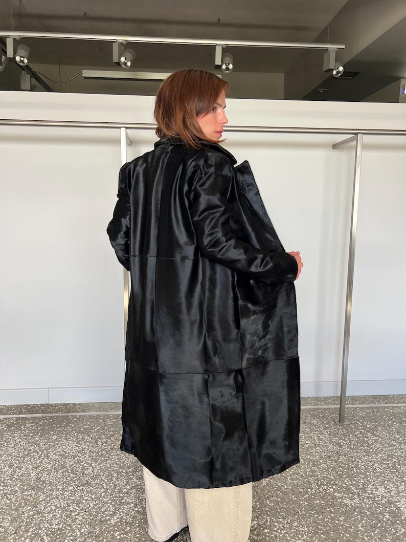 May include: A long, black coat with a shiny, textured appearance. The coat has a tailored fit and a classic design, with a collar and long sleeves. The coat is worn over light-colored pants. The background is a white wall and a clothing rack.