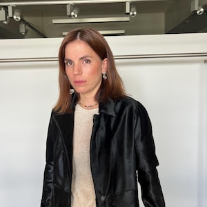 May include: A woman wearing a black coat over a light-colored top and light-colored pants. The coat has a classic collar and button closure. She is also wearing a gold necklace and earrings.