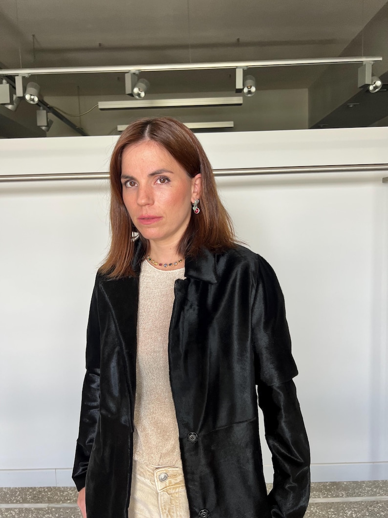 May include: A woman wearing a black coat over a light-colored top and light-colored pants. The coat has a classic collar and button closure. She is also wearing a gold necklace and earrings.