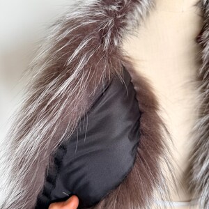 May include: Close-up of a luxurious fur collar with a silver and brown color palette. The collar features long, soft fur and a black lining. The garment is displayed on a mannequin, showcasing the texture and design.