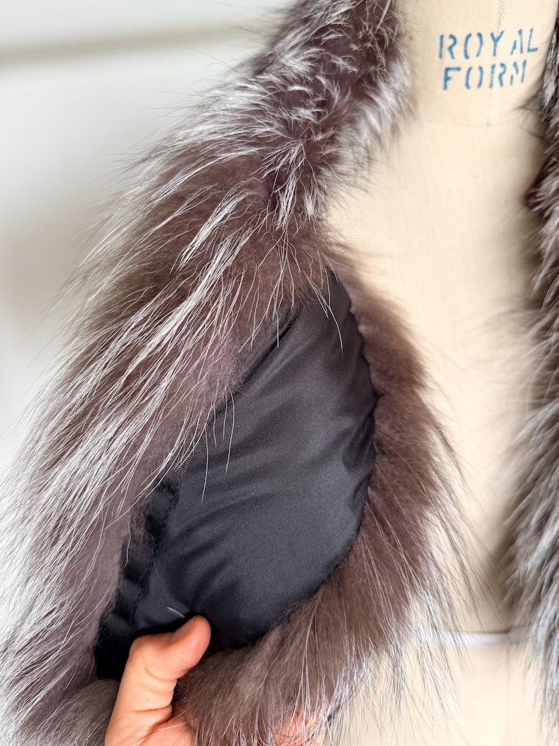 May include: Close-up of a luxurious fur collar with a silver and brown color palette. The collar features long, soft fur and a black lining. The garment is displayed on a mannequin, showcasing the texture and design.