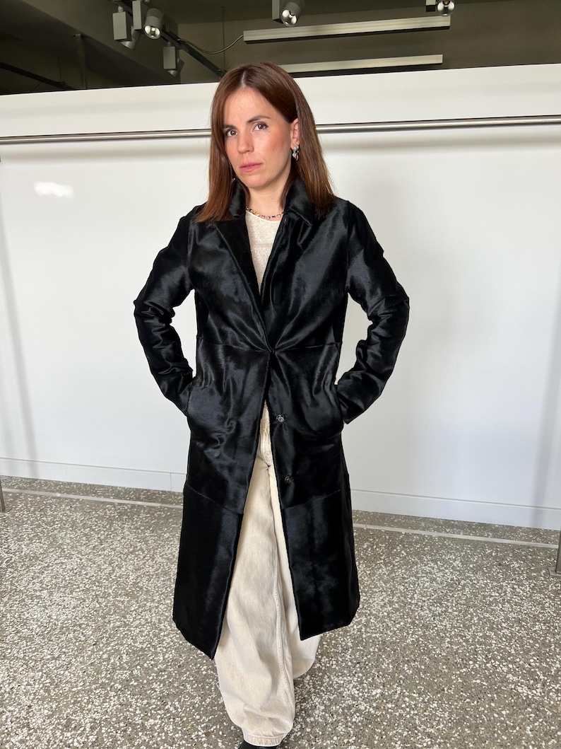 May include: A long, black coat with a shiny, textured appearance. The coat has a classic lapel collar, a single-button closure, and pockets. The person is wearing a cream-colored top and pants. The coat appears to be made of a luxurious material.