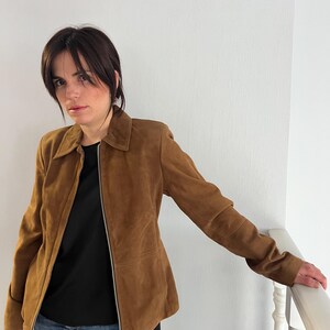 May include: A brown suede jacket with a zip fastening. The jacket features a collared neckline and long sleeves. The person in the image is wearing a black top and blue jeans. The jacket is a medium brown colour.