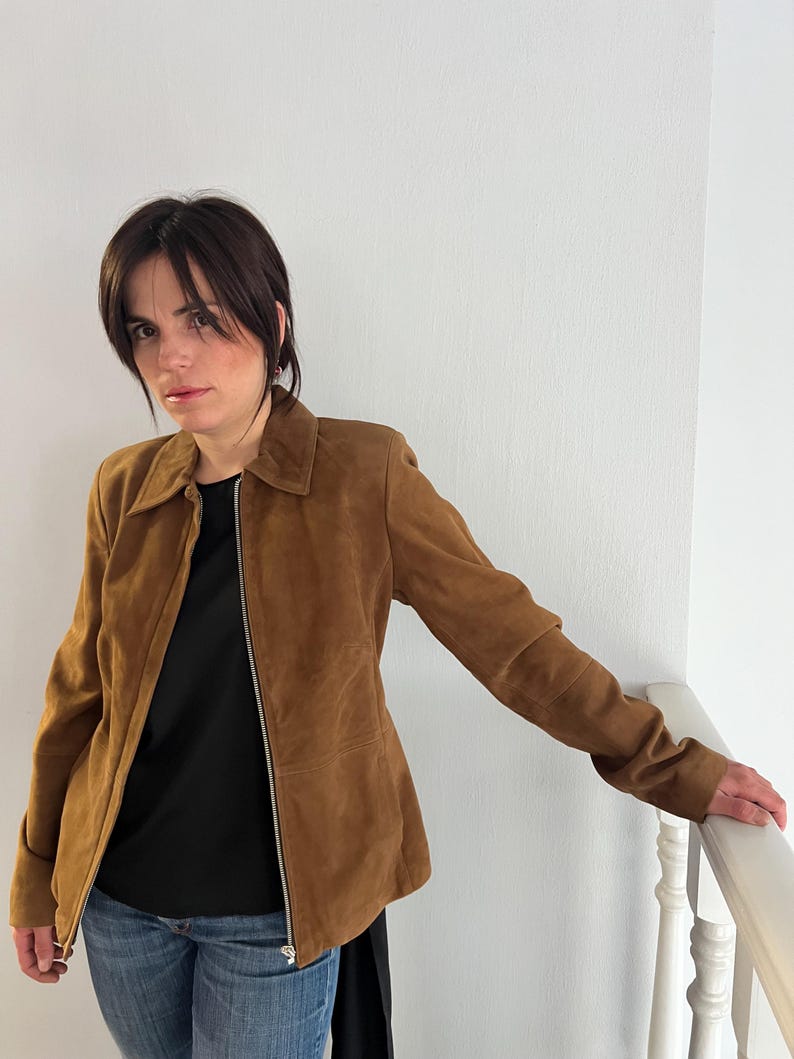 May include: A brown suede jacket with a zip fastening. The jacket features a collared neckline and long sleeves. The person in the image is wearing a black top and blue jeans. The jacket is a medium brown colour.