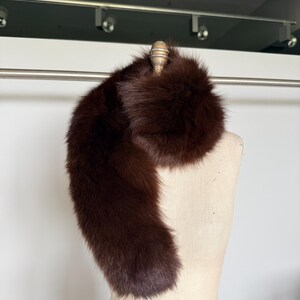 May include: A luxurious brown fur stole, draped over a mannequin. The stole is a rich, dark brown color, with a soft, plush texture. The fur appears to be in excellent condition, with a natural sheen. The stole is designed to be worn around the neck and shoulders.