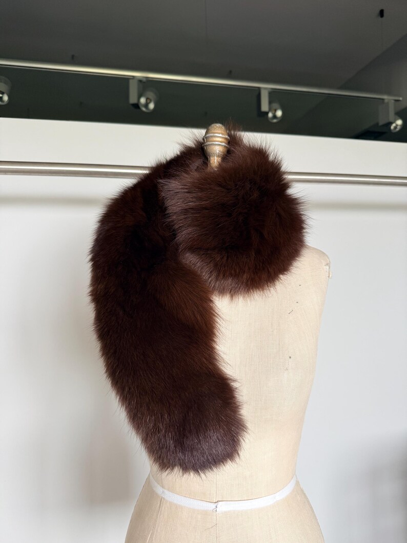 May include: A luxurious brown fur stole, draped over a mannequin. The stole is a rich, dark brown color, with a soft, plush texture. The fur appears to be in excellent condition, with a natural sheen. The stole is designed to be worn around the neck and shoulders.