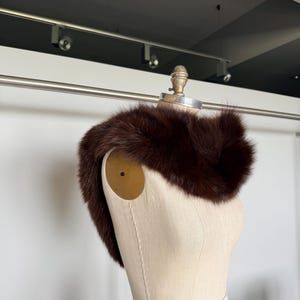 May include: A dark brown fur collar displayed on a mannequin. The fur appears soft and plush, with a rich, deep color. The mannequin is a neutral beige, and the background is a simple white wall.