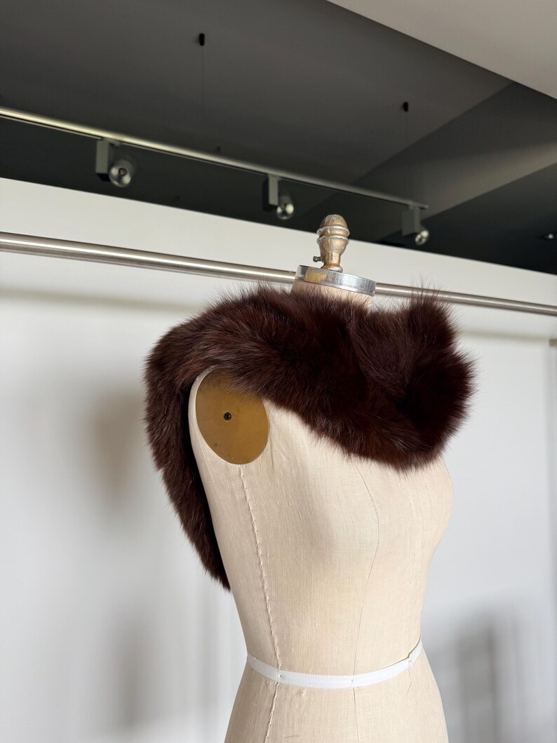 May include: A dark brown fur collar displayed on a mannequin. The fur appears soft and plush, with a rich, deep color. The mannequin is a neutral beige, and the background is a simple white wall.